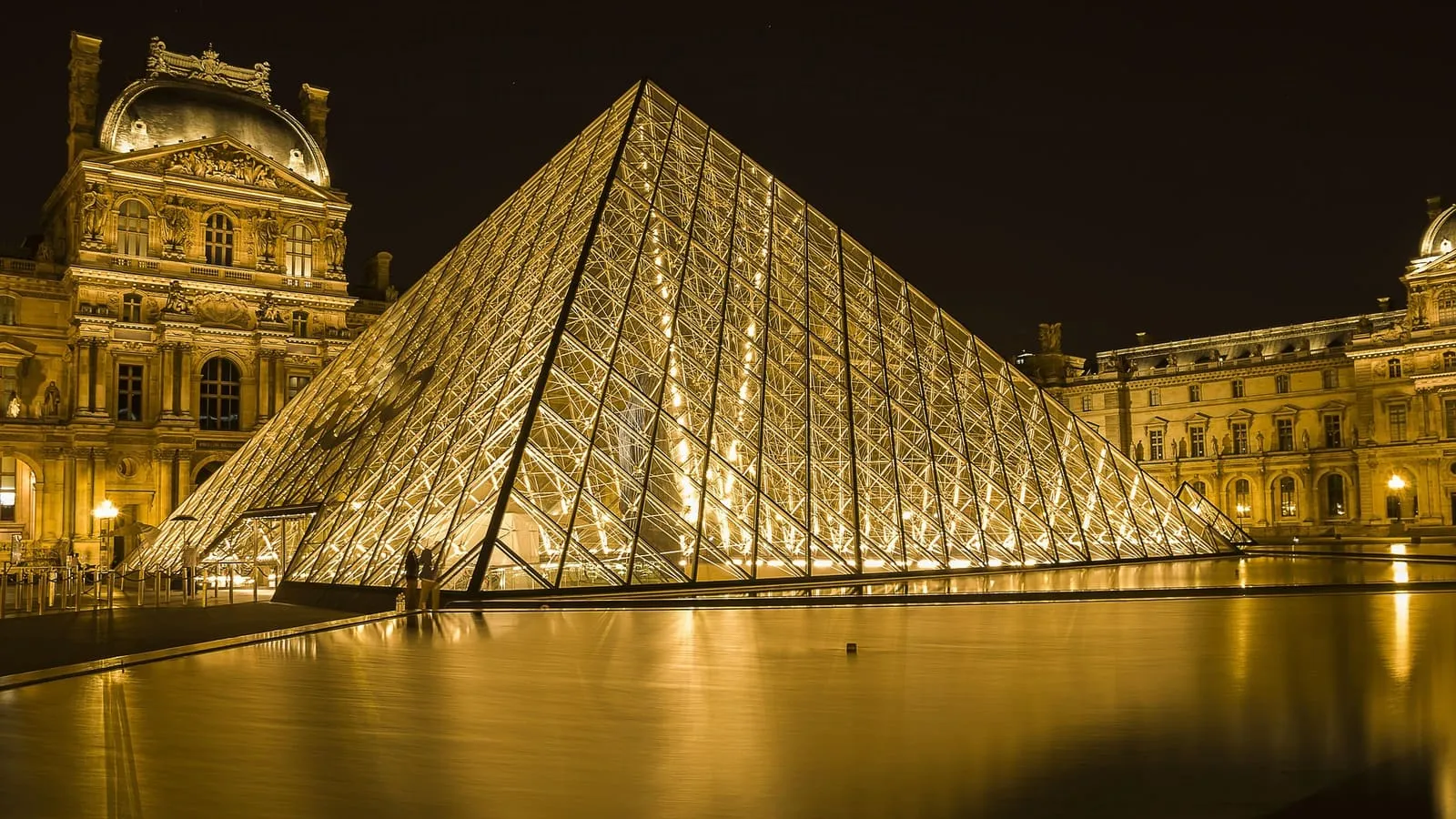 The Louvre pyramid glowing at night, reflected in the fountain basin